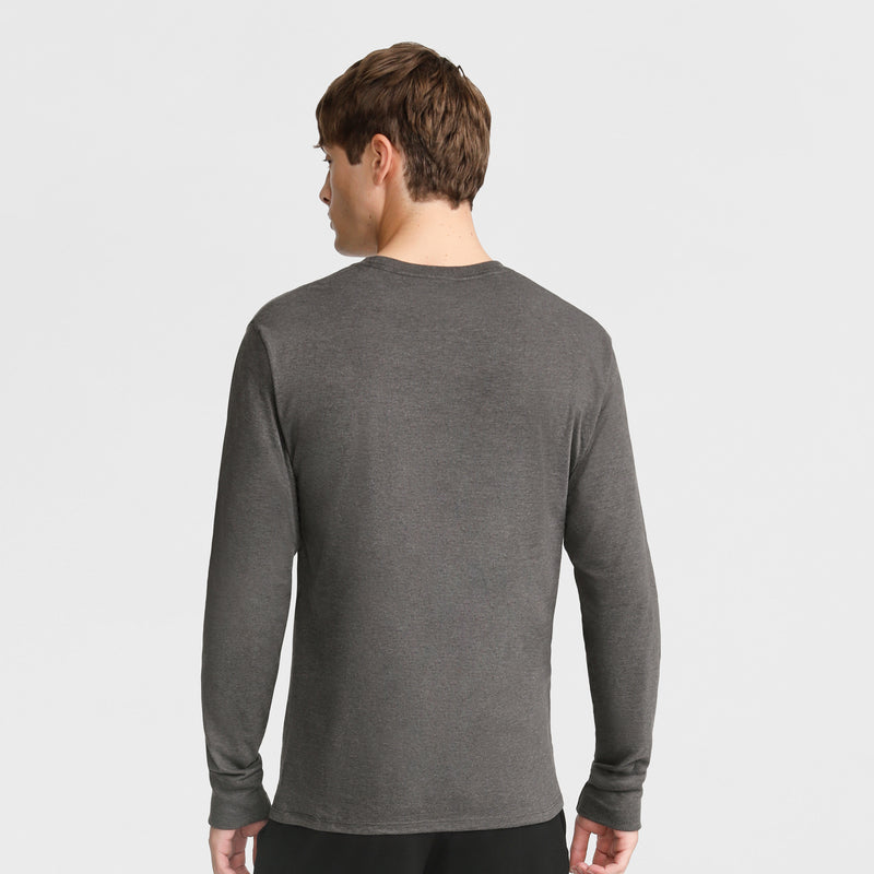 A man with short brown hair stands with his back to the camera, wearing a Champion Classic Graphic Long-Sleeve T-Shirt with a Script Logo in dark gray and black pants against a plain white background.