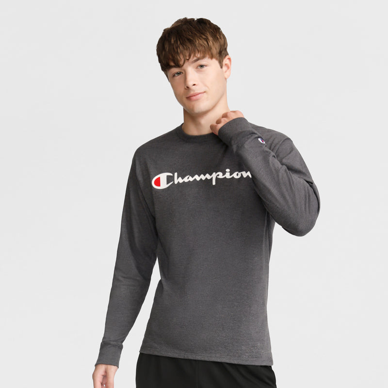 A young man with short brown hair wears a Champion Classic Graphic Long-Sleeve T-Shirt with a script logo and black pants, standing against a plain light gray background. He holds the collar of his soft cotton shirt while facing the camera.