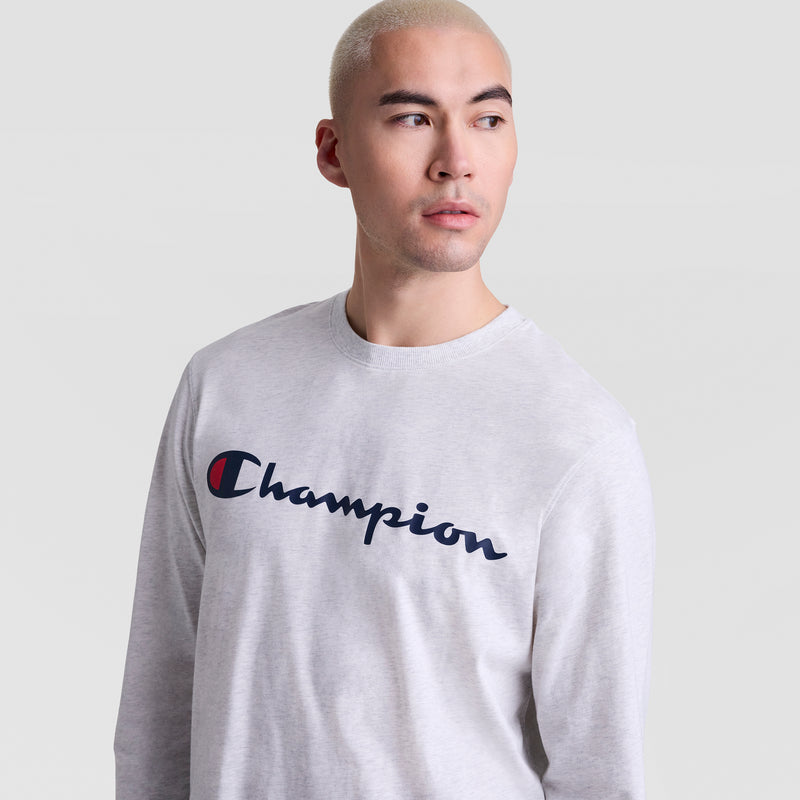 A person with short, light blond hair wears a light gray Champion Classic Graphic Long-Sleeve T-Shirt with a blue script logo, looking slightly to the side against a plain background.