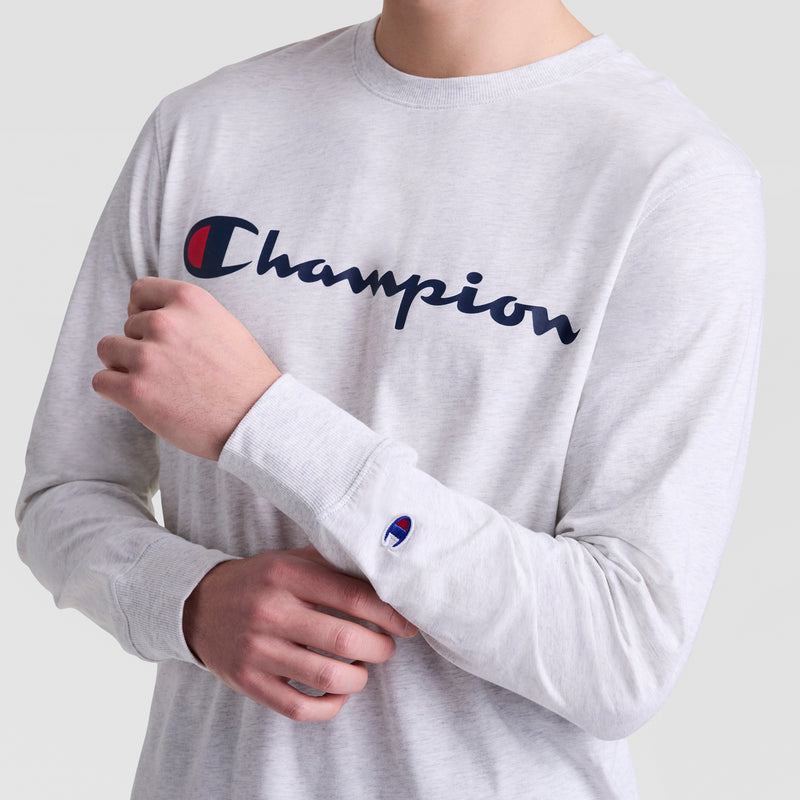 A person wearing a light gray Champion Classic Graphic Long-Sleeve T-Shirt with the iconic script logo across the chest and sleeve stands against a plain background in this soft cotton tee.