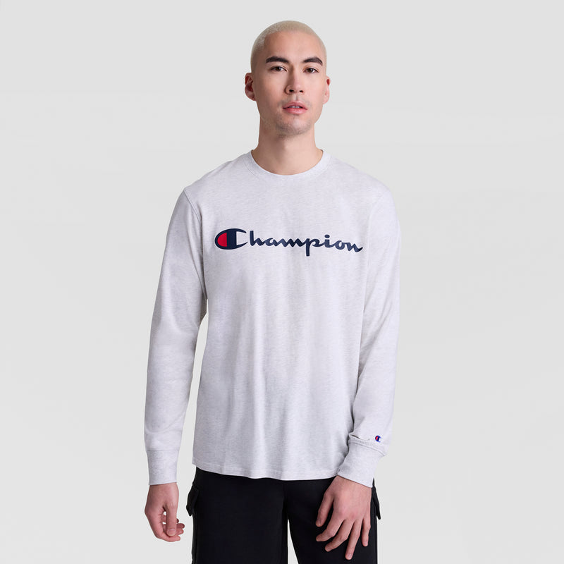 A person with short blond hair wears a Champion Classic Graphic Long-Sleeve T-Shirt with a script logo in light gray and black pants, standing against a plain light gray background.