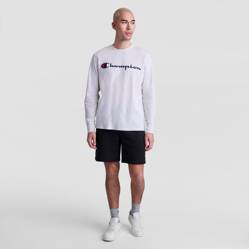 A person with short blond hair wears a white Champion Classic Graphic Long-Sleeve T-Shirt with Script Logo, black shorts, gray socks, and white sneakers while standing against a plain light gray background.