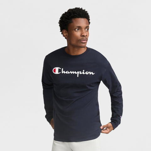 A man wears a navy Classic Graphic Long-Sleeve T-Shirt with the Champion script logo in white, standing against a plain white background, looking slightly left with his hands by his sides.