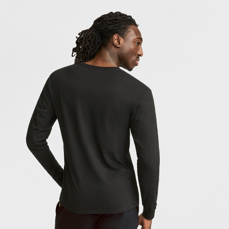 A person with long, dark, braided hair faces away, wearing a Champion Classic Graphic Long-Sleeve T-Shirt with script logo and black pants against a plain light background.