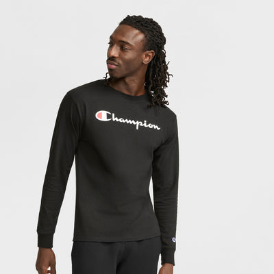 A man with long dreadlocks wears a black Champion Classic Graphic Long-Sleeve T-Shirt with a script logo and black pants, looking to the side with a neutral expression against a plain white background.