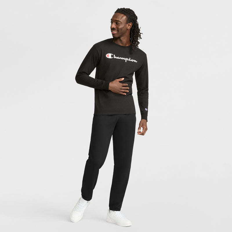 A man smiles while wearing Champion’s Classic Graphic Long-Sleeve T-Shirt with a script logo, paired with black pants and white sneakers. He stands against a plain white background, one hand resting on his stomach as he looks to the side.