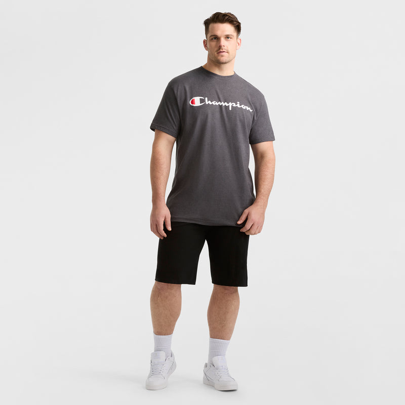 A man models a dark gray Champion Classic Graphic T-Shirt, Script Logo - Big & Tall, paired with black shorts, white socks, and white sneakers against a plain white background for a classic streetwear look.