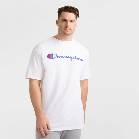A man with short brown hair wears a white Champion Classic Graphic T-Shirt, Script Logo - Big & Tall, featuring the brand’s iconic blue and red script logo, paired with light gray pants against a plain white background.