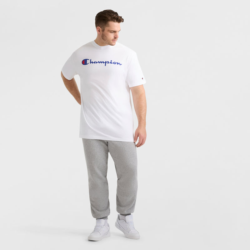 A man in a white Champion Classic Graphic T-Shirt with Script Logo (Big & Tall) stands against a plain light background, paired with gray sweatpants and white sneakers, embodying classic streetwear as he looks down and to the side.