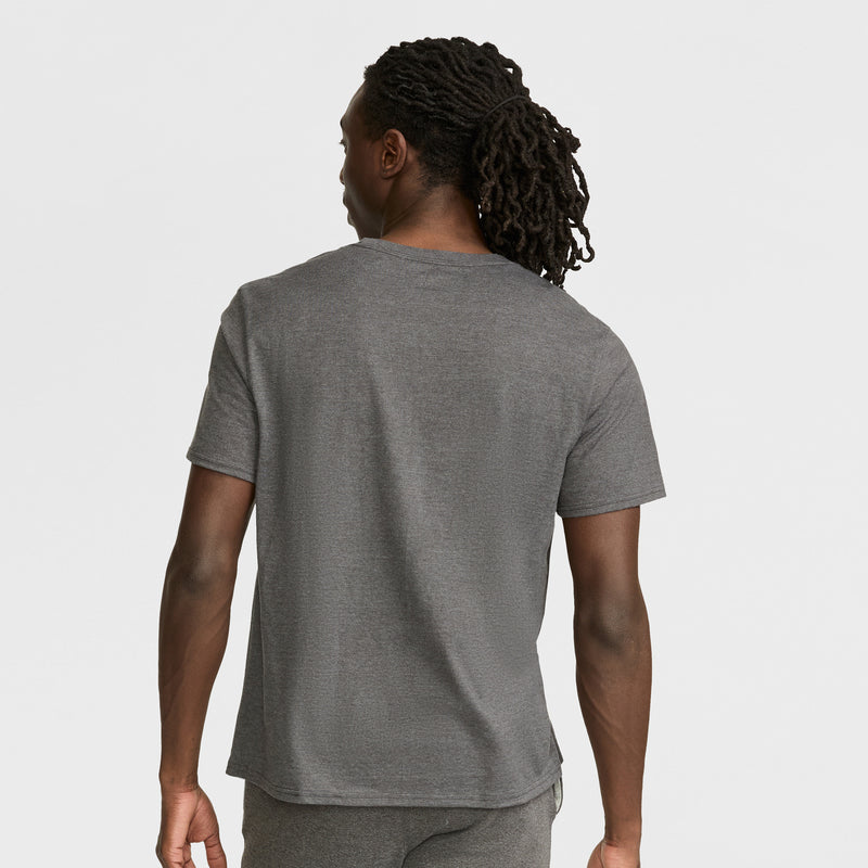 A person with long, dark, braided hair stands with their back to the camera, wearing a Champion Classic Graphic T-Shirt with a script logo and gray pants in a casual streetwear style against a white background.