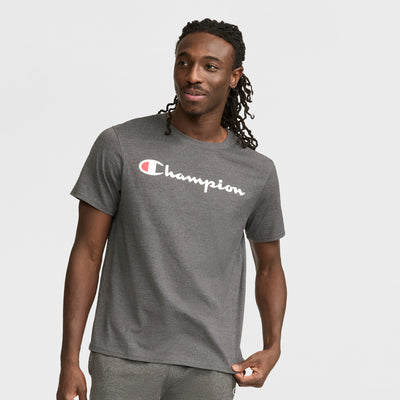 A man with long braided hair wears a Champion Classic Graphic T-Shirt with a script logo, smiling and looking to the side, effortlessly showcasing timeless streetwear style against a plain light background.