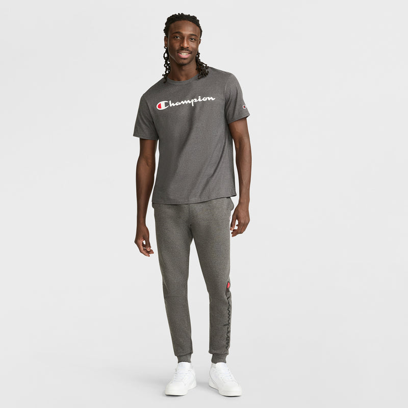 A man smiles, wearing a Champion Classic Graphic T-Shirt with the script logo and matching joggers, paired with white sneakers, presenting a relaxed streetwear look against a plain light background.