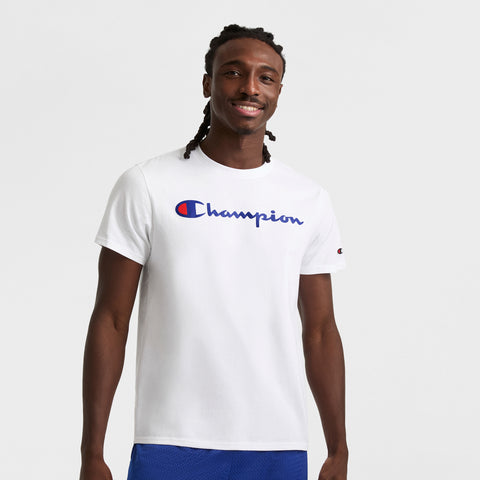 A man with long braided hair smiles in a white Champion Classic Graphic T-Shirt with a script logo and blue shorts, standing against a plain white background—a relaxed streetwear vibe.