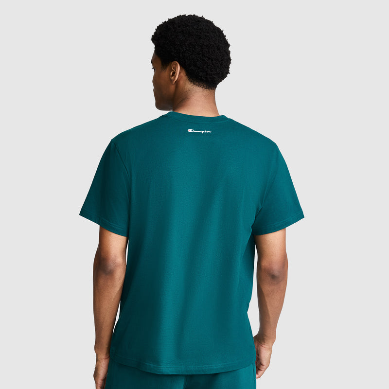 A person stands facing away, wearing a teal Classic Graphic T-Shirt, Script Logo by Champion with matching shorts. The 100% cotton men’s t-shirt features a small, white Champion script logo on the upper back. The background is plain light gray.