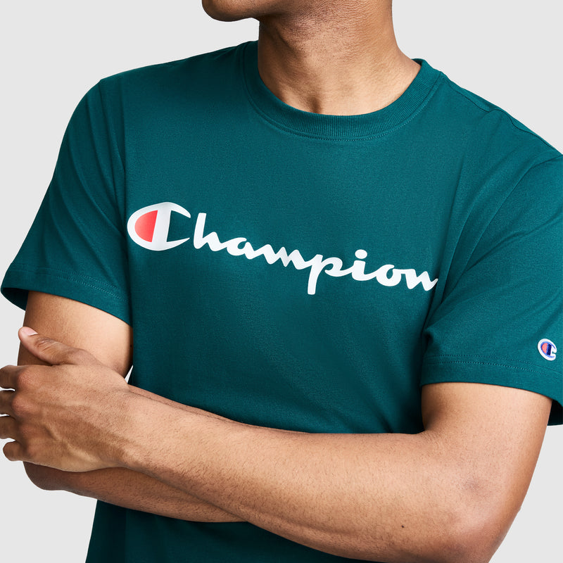 A person wearing the Champion Classic Graphic T-Shirt, Script Logo in green stands with arms crossed, showcasing the bold white script logo across the chest. The 100% cotton t-shirt pops against a plain light gray background.