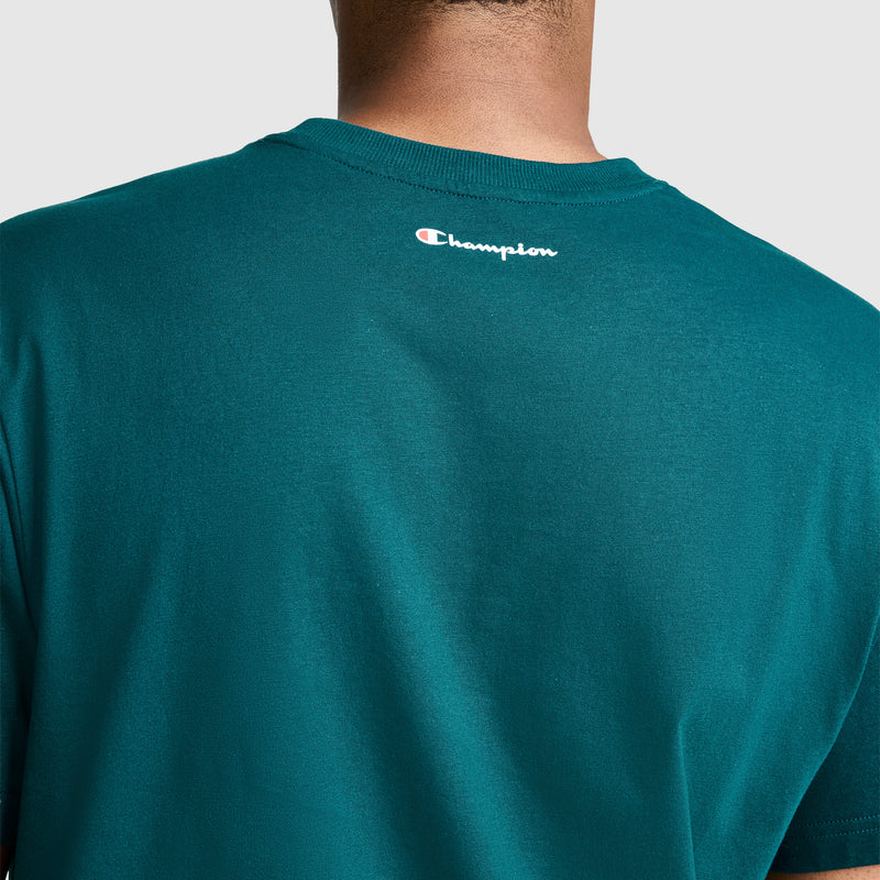 A person is shown from the back wearing a teal Champion Classic Graphic T-Shirt, Script Logo, made from 100% cotton. The small Champion logo appears near the neckline of the shirt.