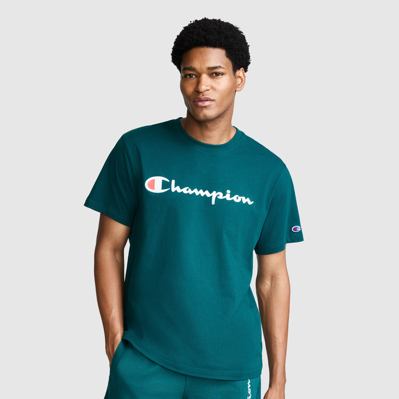 A man wearing a teal Champion Classic Graphic T-Shirt with the Script Logo and matching shorts stands against a plain light gray background with one hand in his pocket.
