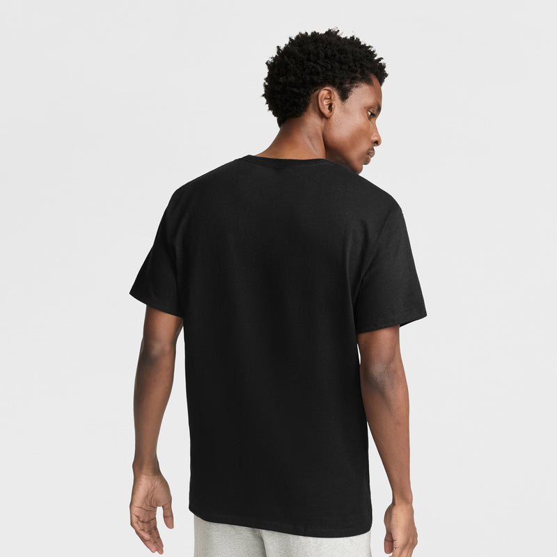 A person with short curly hair is standing and facing away, wearing a Champion Classic Graphic T-Shirt with Script Logo in black and light gray pants against a white background.