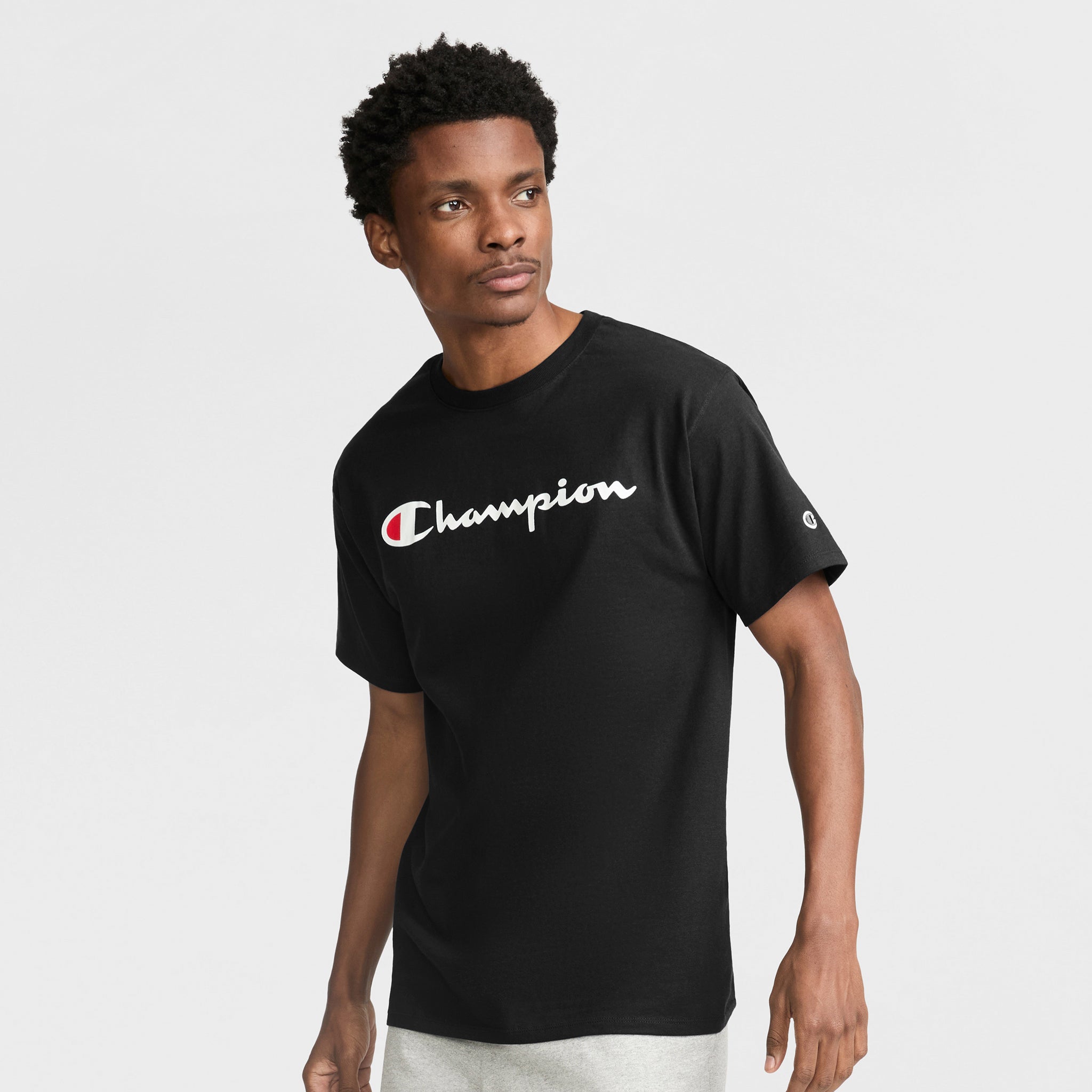 A man in a black Champion Classic Graphic T-Shirt with the iconic script logo stands against a plain light background, gazing slightly to the side.