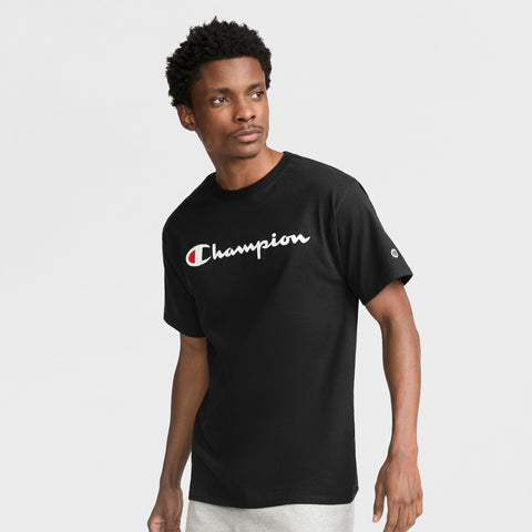A man in a black Champion Classic Graphic T-Shirt with the iconic script logo stands against a plain light background, gazing slightly to the side.