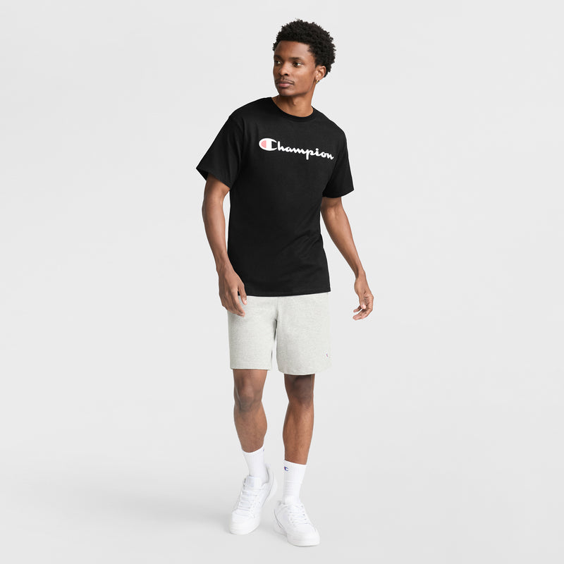 A man wears a black Champion Classic Graphic T-Shirt with the script logo, paired with light gray shorts, white socks, and white sneakers as he stands against a plain white background, gazing slightly to his left in casual streetwear style.