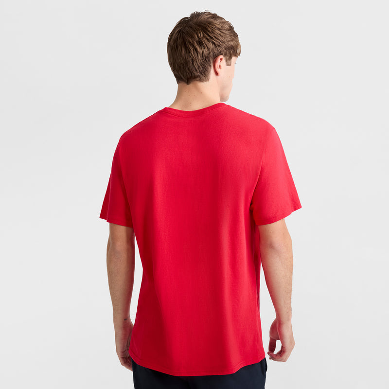 A man with short brown hair faces away from the camera, wearing a Champion Classic Graphic T-Shirt with Script Logo in red, paired with dark pants against a light background.