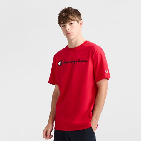 A young man with short brown hair models the Champion Classic Graphic T-Shirt, Script Logo in bright red and black pants, exuding laidback streetwear style against a plain light background with his left hand in his pocket.