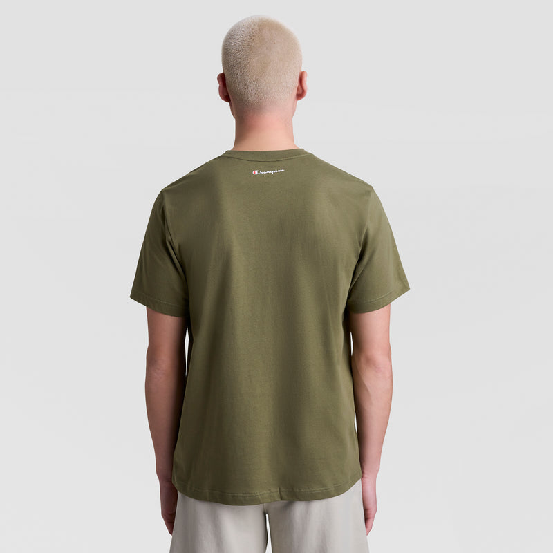 A person with short, bleached blond hair is shown from behind in a Champion Classic Graphic T-Shirt with the Script Logo in olive green and light shorts, standing against a plain light gray background.