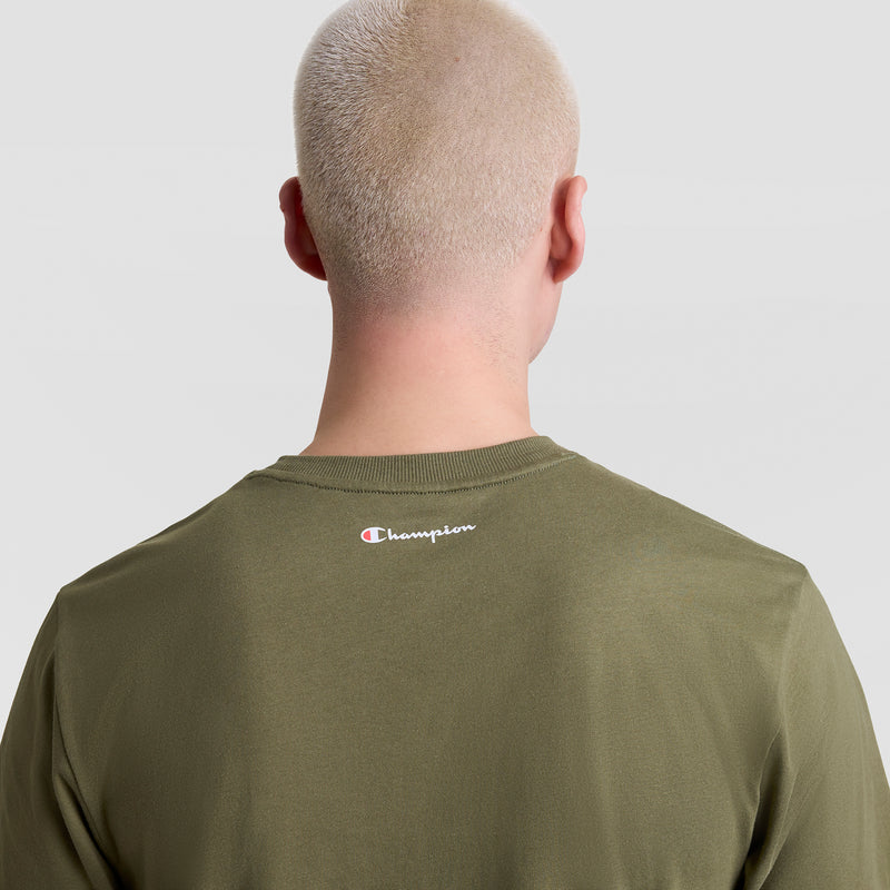 A person with short, light blonde hair is seen from behind wearing a Champion Classic Graphic T-Shirt with the script logo near the neckline, set against a plain light background.