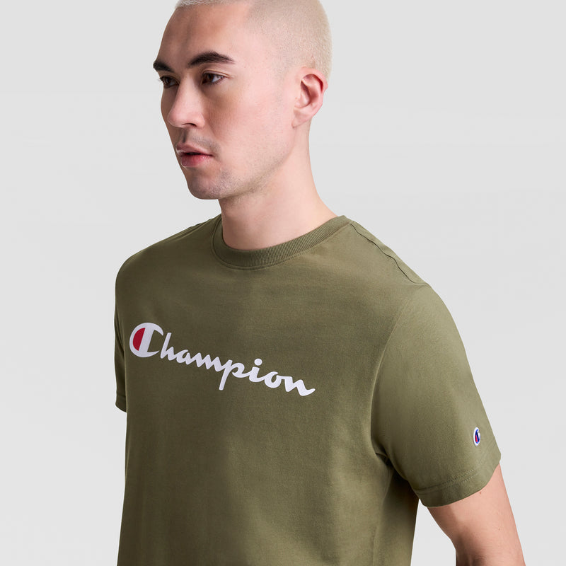 A person with short blonde hair wears a Champion Classic Graphic T-Shirt, Script Logo, in olive green with the white and red logo on the chest, standing against a plain light gray background.