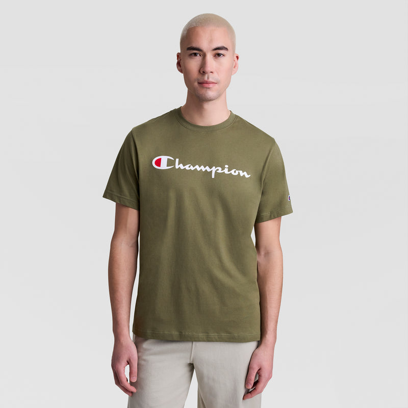 A person with short blonde hair wears a Champion Classic Graphic T-Shirt with the script logo in olive green, styled as casual streetwear with light pants, standing against a plain light gray background.