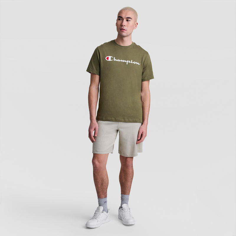 A person with a shaved head wears a Champion Classic Graphic T-Shirt with Script Logo in olive green, paired with beige shorts, gray socks, and white sneakers, standing against a plain white background.