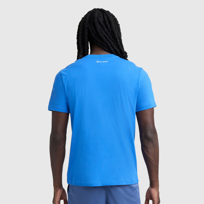 A person with long, twisted hair is seen from the back wearing Champion’s Classic Graphic T-Shirt, Script Logo in bright blue and matching shorts—perfect streetwear style against a plain light gray background.