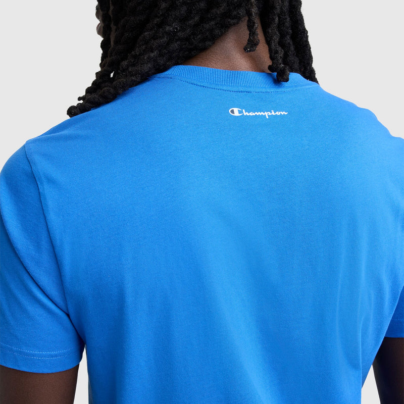 A person with long braids is seen from behind in a bright blue Champion Classic Graphic T-Shirt, Script Logo—a standout streetwear piece featuring the white script logo on the upper back, set against a plain light background.