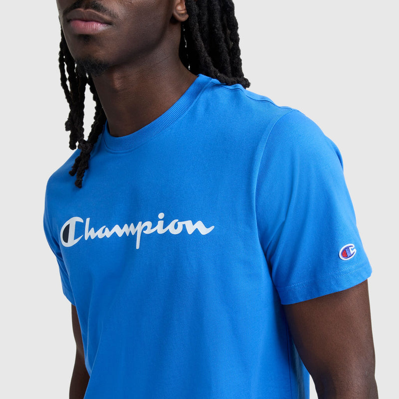 A person with long, dark, braided hair wears a bright blue Champion Classic Graphic T-Shirt, Script Logo—a streetwear staple featuring a bold white logo across the chest and a small embroidered sleeve logo.