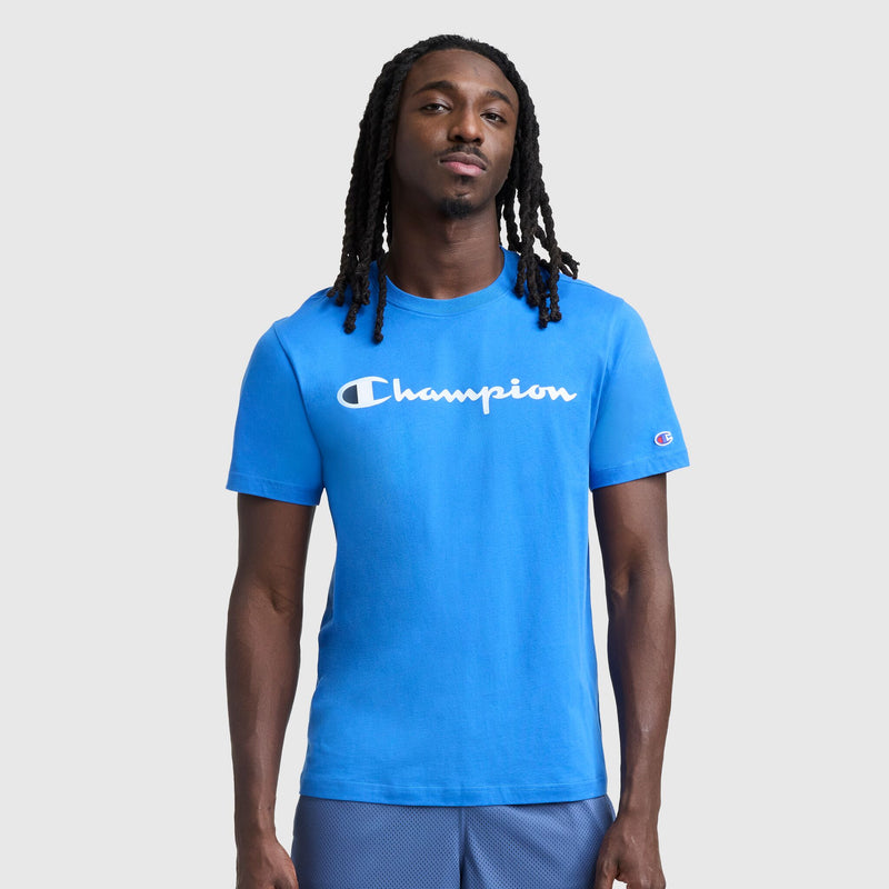 A man with long dreadlocks wears a bright blue Champion Classic Graphic T-Shirt, Script Logo, embodying iconic streetwear style against a plain light gray background.