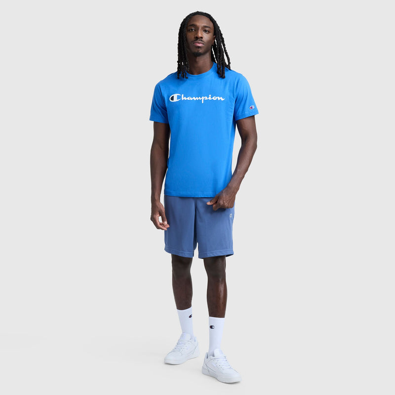 A man stands facing forward against a light gray background, wearing a blue Champion Classic Graphic T-Shirt with the Script Logo, styled as streetwear with gray shorts, white socks, and white sneakers.