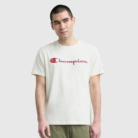 A man wears the Champion Classic Graphic T-Shirt with a red script logo on the front, standing against a plain light gray background and looking to the side for a timeless streetwear vibe.