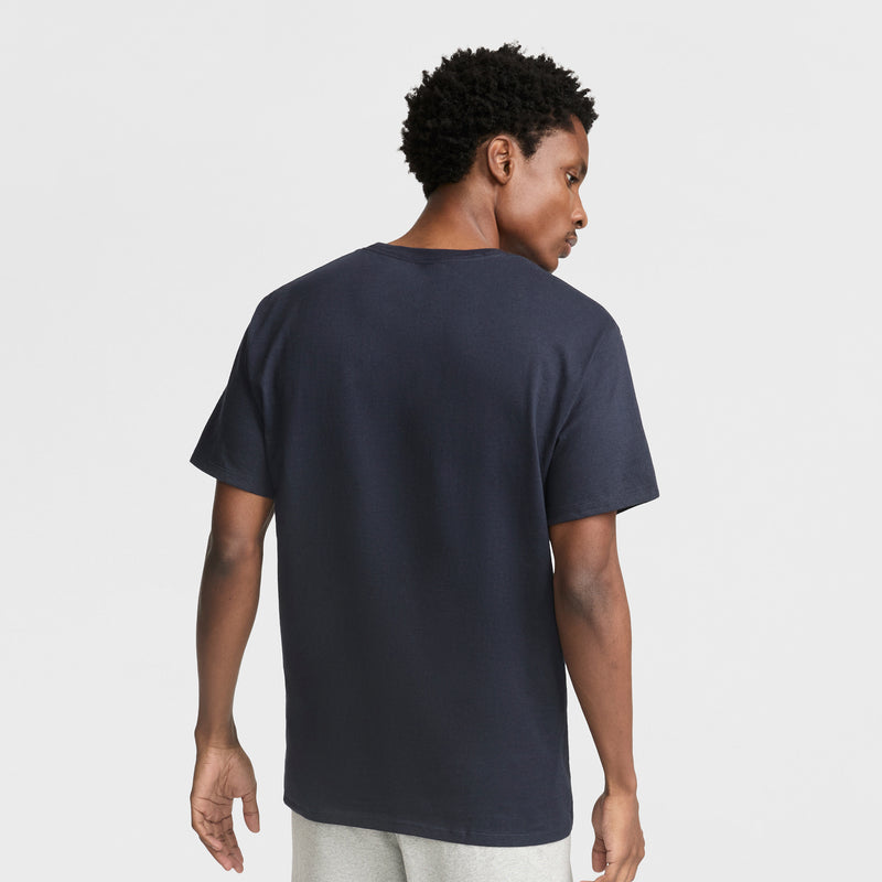 A man with short curly hair stands with his back to the camera, highlighting the Champion Classic Graphic T-Shirt with Script Logo and light gray pants, styled in laid-back streetwear against a white backdrop.