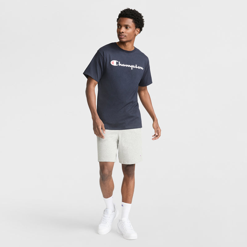 A man stands against a white backdrop in a navy Champion Classic Graphic T-Shirt with Script Logo, paired with light gray shorts, white crew socks, and sneakers—capturing an easy streetwear look as he glances sideways with one arm slightly raised.