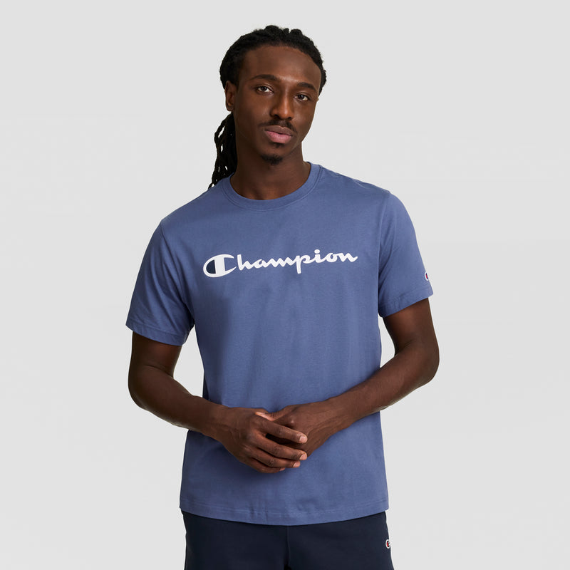 A man with long braided hair wears a blue Champion Classic Graphic T-Shirt with the Script Logo, standing against a plain light gray background, facing the camera with a neutral expression and hands loosely clasped.