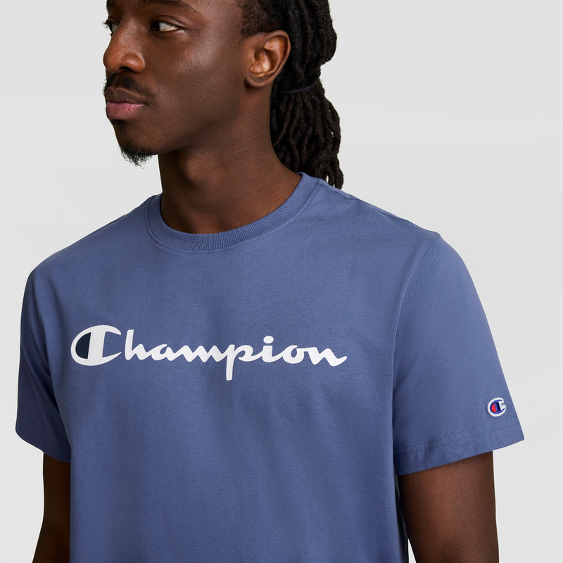 A man with long braided hair wears the Champion Classic Graphic T-Shirt, Script Logo, featuring the white Champion script across the chest and a sleeve patch, perfect for streetwear style against a plain light background.
