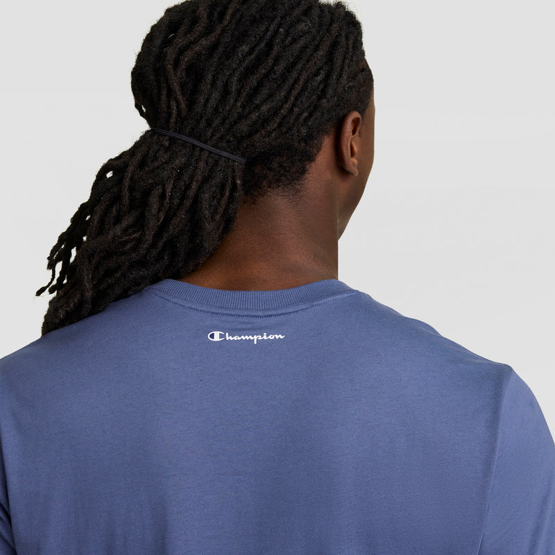 A person with long, dark dreadlocks tied back is seen from behind wearing Champion's Classic Graphic T-Shirt, Script Logo in blue, featuring the white logo on the upper back. The plain light background emphasizes this signature streetwear style.