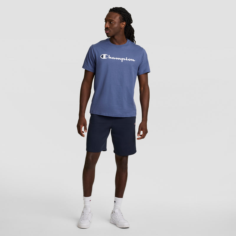 A man in a Champion Classic Graphic T-Shirt with the script logo, navy shorts, white socks, and sneakers stands against a plain background, embracing streetwear style as he looks left with his arms relaxed at his sides.