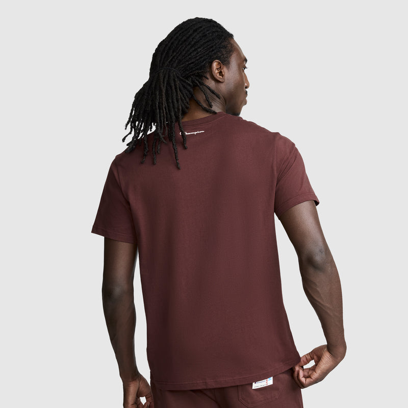 A person with long, dark hair in a ponytail is seen from behind, standing against a plain light gray background. They are wearing the Champion Classic Graphic T-Shirt with Script Logo in maroon and matching pants, creating an effortlessly casual streetwear look.