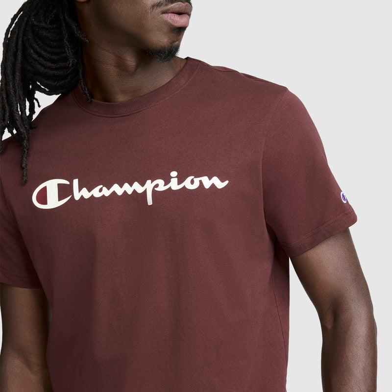 A person wearing the Champion Classic Graphic T-Shirt, Script Logo in maroon—a men’s tee featuring the iconic white Champion script logo across the chest—shown from shoulders to waist and facing slightly to the side, capturing a modern streetwear staple.