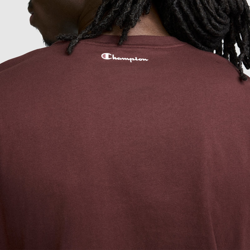A person with dark, twisted hair wears the Champion Classic Graphic T-Shirt, Script Logo—a maroon men’s tee that perfectly embodies streetwear style. The back of this shirt displays a small white script logo near the neckline.