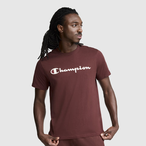 A man with long dreadlocks stands against a plain light gray background in true streetwear style, wearing the maroon Champion Classic Graphic T-Shirt with Script Logo and matching pants, looking to his left.