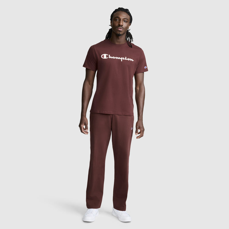 A man stands facing forward, showcasing a Champion Classic Graphic T-Shirt with the signature Script Logo in maroon, paired with matching pants and white sneakers for a modern streetwear look against a plain light gray background.
