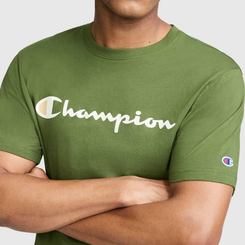 A person wearing the Champion Classic Graphic T-Shirt, Script Logo in green, featuring the signature Champion script across the chest and a small emblem on the sleeve. Channeling classic streetwear style, their arms are crossed and their face is partially obscured.